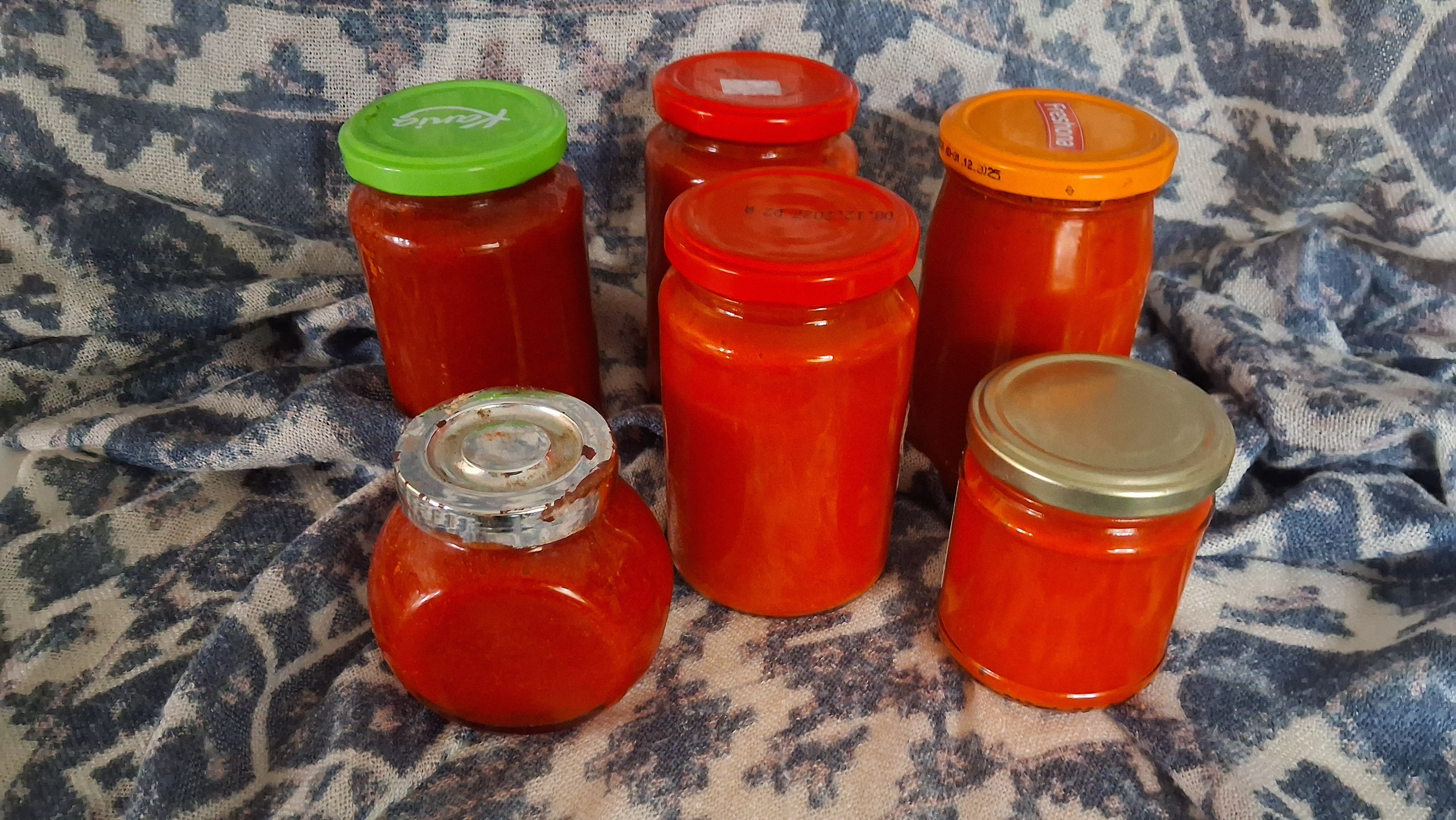 Assorted glass jars filled with red tomato sauce on a blue patterned fabric background, with colorful lids (green, red, orange, metal).