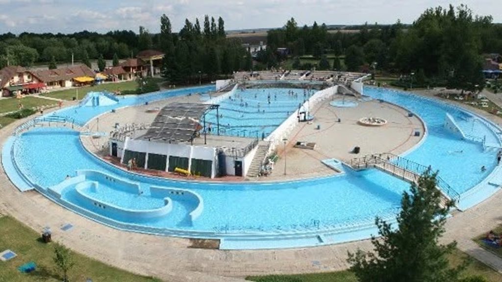 Aerial view of a circular outdoor water park with blue pools, slides, and surrounding greenery and buildings.