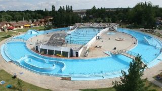 Aerial view of a circular outdoor water park with blue pools, slides, and surrounding greenery and buildings.