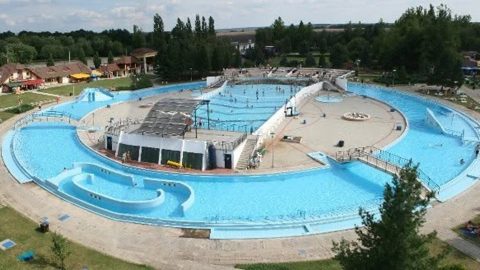 Aerial view of a circular outdoor water park with blue pools, slides, and surrounding greenery and buildings.