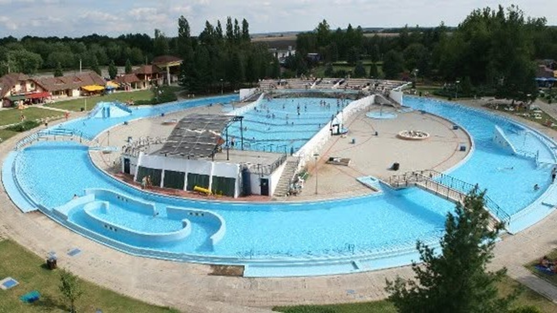 Aerial view of a circular outdoor water park with blue pools, slides, and surrounding greenery and buildings.