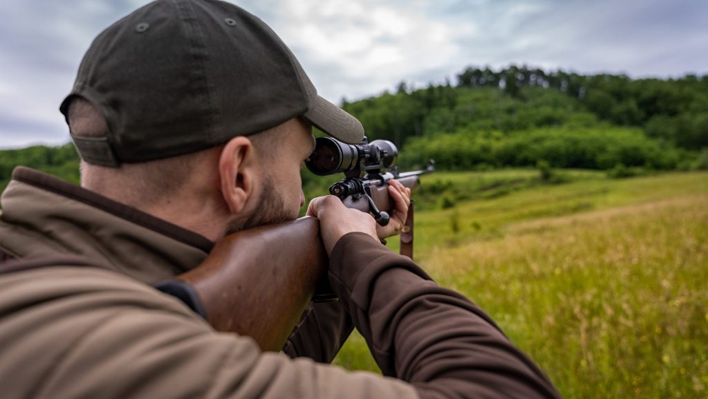 Man wearing a dark cap and brown jacket, aiming a scoped rifle in a grassy field.
