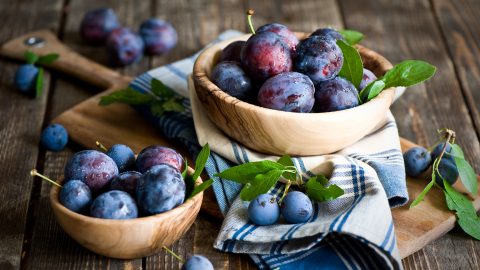 Wooden bowls of ripe blue plums with green leaves on a rustic wooden table.