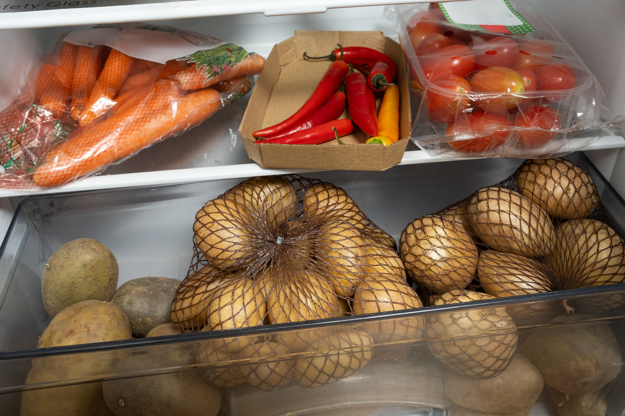 Carton and bags of fresh vegetables in a fridge: carrots (left), red and yellow peppers in a box (center), and tomatoes in a plastic container (right) with netted potatoes below.