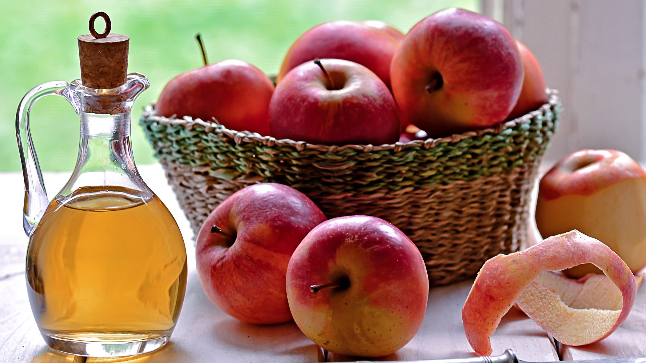 Glass bottle of golden oil with a cork stopper beside a basket of red apples and apple peels on a light wooden surface