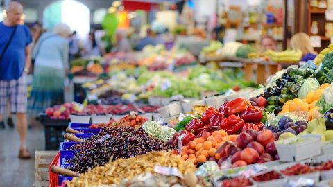 Vibrant fruit and vegetable market stall with colorful produce and shoppers in the background.